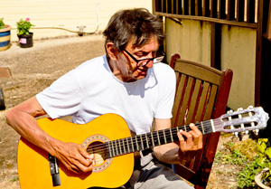 One of the people we support, sitting outside on a summer's day, playing his guitar. He is wearing a white t-shirt and sitting on a brown chair.