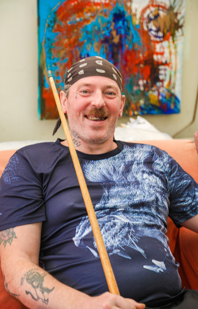 One of the people we support, sitting in our pool room, grinning. He is wearing a black t-shirt with a white image on it and a black and white bandanna tied around his head. He has a moustache and looks very happy. In in right hand he is a holding a pool cue. There is a colourful painting on the wall behind him.