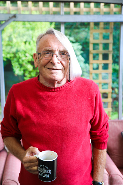 One of the people we support, holding a cup of tea in his right hand. He has white hair, cut very short on one side and long on the other and is wearing a red jumper. In the background you can see a trellis and the green of bushes and trees.